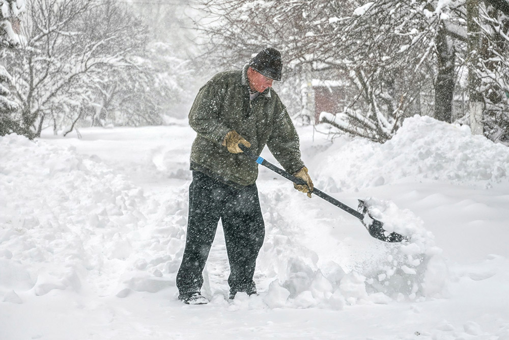 An older man in a dark cloak and cap actively shoveling snow during heavy snowfall