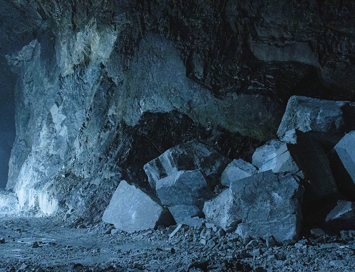 Collapsed cave entrance surrounded by dust, warning barriers, and investigators in protective gear.